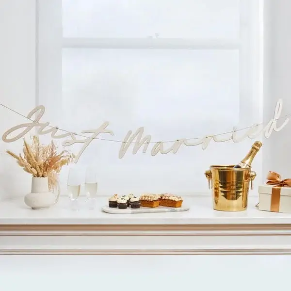 A Just Married banner draped across a bright white windowsill styled with champagne flutes, pastries, dried flowers, a gold ice bucket with champagne and a wrapped gift.